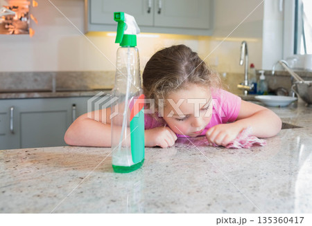 Child cleaning granite countertop in home kitchen with spray bottle and pink cloth, copy space Child cleaning granite countertop in home kitchen with spray bottle and pink cloth, copy space 135360417