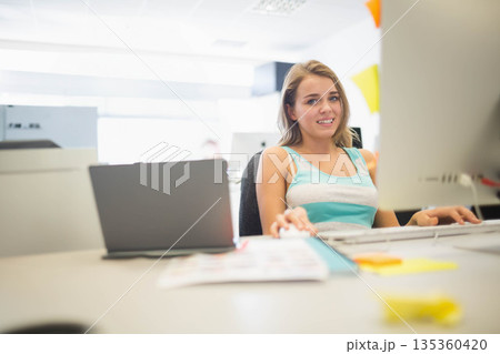 Female office worker working on laptop and monitor at office desk with sticky notes, copy space Female office worker working on laptop and monitor at office desk with sticky notes, copy space 135360420