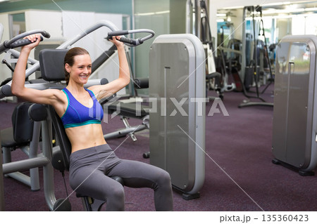 Female gym goer wearing sports bra and leggings using shoulder press machine at gym, copy space Female gym goer wearing sports bra and leggings using shoulder press machine at gym, copy space 135360423