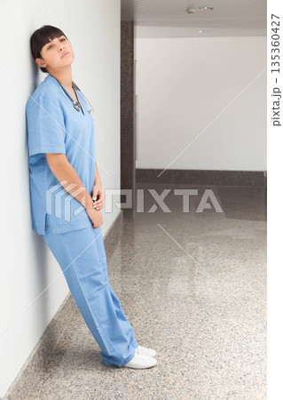 Hispanic woman nurse leaning on wall in hospital hallway wearing scrubs, stethoscope, copy space 135360427