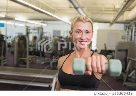Female in sportswear standing before mirror wall holding green hex dumbbell toward camera at gym 135360438
