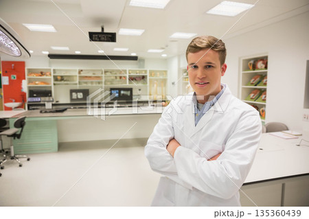 Male researcher wearing lab coat with arms crossed at medical lab near monitors and anatomy models Male researcher wearing lab coat with arms crossed at medical lab near monitors and anatomy models 135360439
