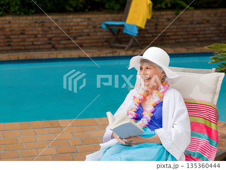 Senior woman wearing sun hat and floral lei reading book on pool patio chair, copy space 135360444