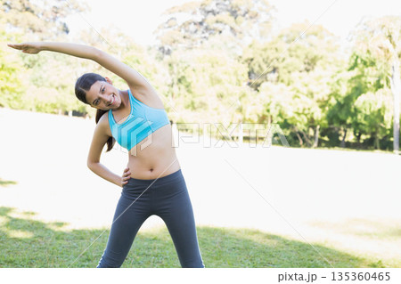 Adult fitness instructor performing side stretch in turquoise bra leggings on park lawn, copy space Adult fitness instructor performing side stretch in turquoise bra leggings on park lawn, copy space 135360465