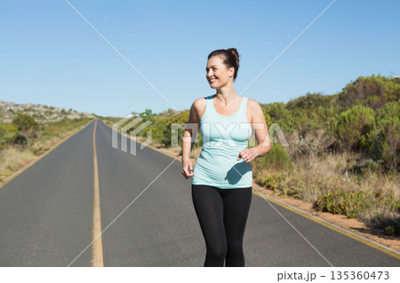 Female jogger jogging along straight two-lane rural road under clear sky with central yellow line Female jogger jogging along straight two-lane rural road under clear sky with central yellow line 135360473