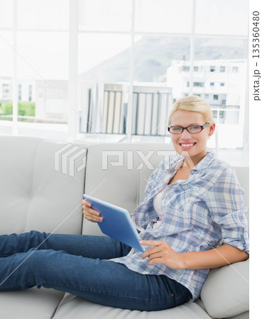 Woman wearing eyeglasses reclining on gray sofa in living room holding blue tablet, copy space Woman wearing eyeglasses reclining on gray sofa in living room holding blue tablet, copy space 135360480