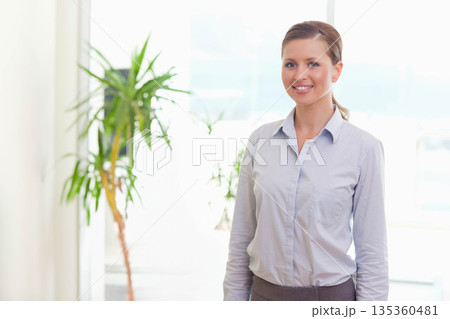 Woman standing by potted plant near large window wearing blue shirt in bright office, copy space 135360481