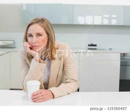 Mature woman sitting at counter in kitchen, wearing blazer and holding coffee cup next to toaster 135360487
