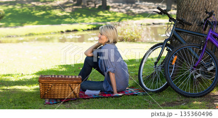Adult woman sitting on plaid blanket by pond in park with picnic basket and bicycles Adult woman sitting on plaid blanket by pond in park with picnic basket and bicycles 135360496