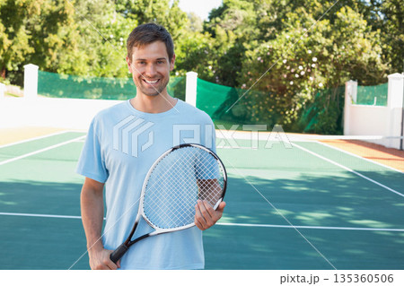 Man smiling and holding tennis racket at chest on sunlit green tennis court by tennis net Man smiling and holding tennis racket at chest on sunlit green tennis court by tennis net 135360506