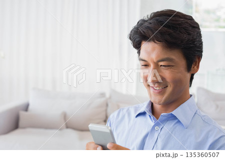Japanese man holding smartphone on sofa at home with cushions, curtains, copy space Japanese man holding smartphone on sofa at home with cushions, curtains, copy space 135360507