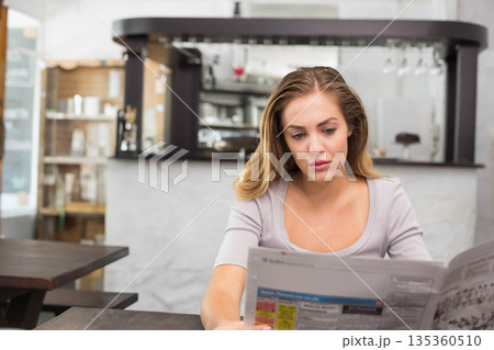 Woman in her twenties reading newspaper at dark wood café table with bar counter, wine glasses 135360510