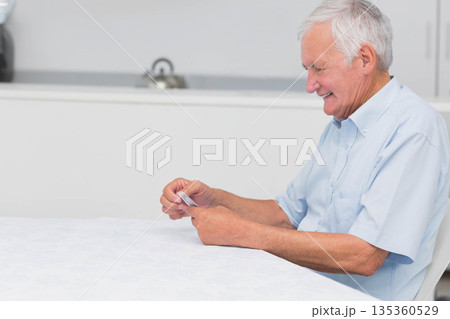 Senior man holding playing cards and smiling at dining table in kitchen with kettle, copy space 135360529