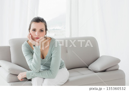 Woman in her thirties sitting on grey leather sofa in living room gazing through sheer curtains Woman in her thirties sitting on grey leather sofa in living room gazing through sheer curtains 135360532
