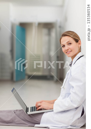 Female doctor sitting in corridor in lab coat and scrubs using laptop stethoscope copy space 135360534