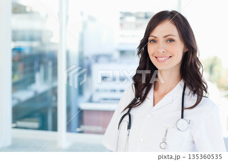 Female doctor standing in clinic office wearing white coat with stethoscope and badge, copy space 135360535