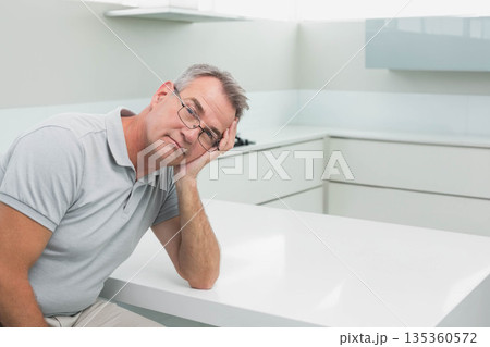 Middle-aged man wearing glasses leaning head on hand sitting at white kitchen island, copy space Middle-aged man wearing glasses leaning head on hand sitting at white kitchen island, copy space 135360572