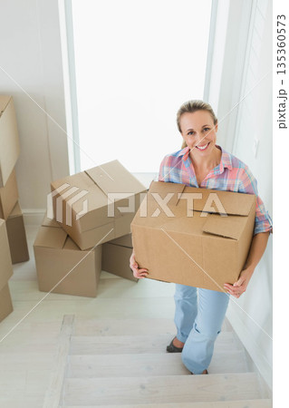 Mid adult woman climbing light-wood stairs inside bright entryway carrying cardboard moving box 135360573