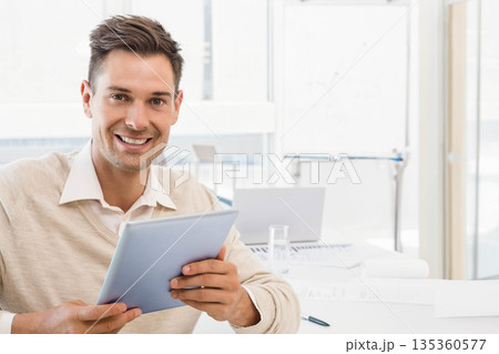 Mid adult man holding tablet, smiling toward camera at office desk with whiteboard, copy space 135360577