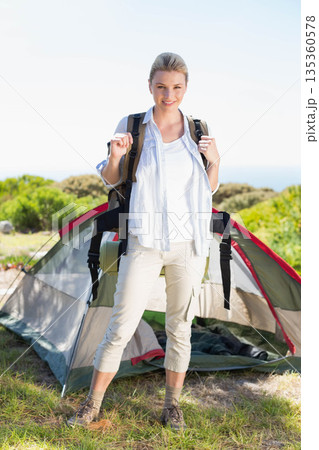 Woman gripping backpack straps, standing on grassy clearing in front of open tent with sleeping mat 135360578