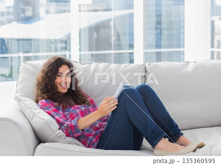 Woman reclining on light-colored sofa in modern living room holding tablet and showing city skyline Woman reclining on light-colored sofa in modern living room holding tablet and showing city skyline 135360595