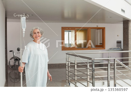Senior woman holding IV pole in hospital corridor next to wheelchair, wearing gown 135360597