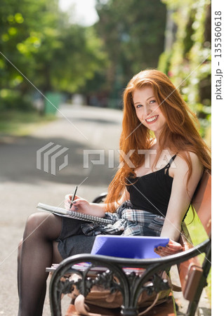 Woman writing notes in notebook on bench in park with pen and blue folder, copy space Woman writing notes in notebook on bench in park with pen and blue folder, copy space 135360618