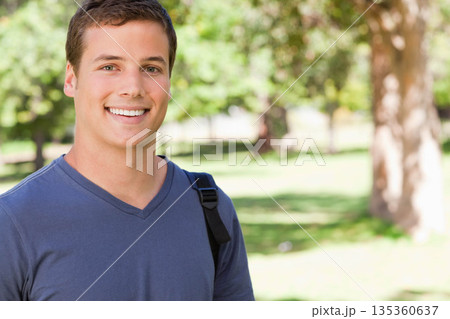 Male student standing smiling in green park wearing blue V-neck T-shirt backpack strap, copy space 135360637