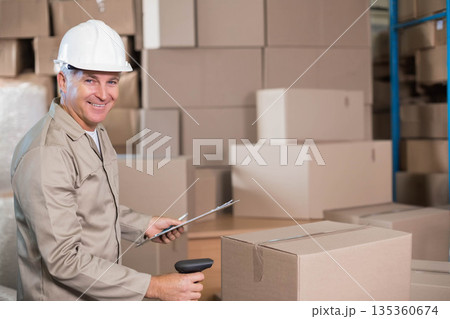 Middle-aged man scanning boxes with scanner and logging items on clipboard in warehouse, copy space 135360674