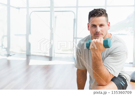 Man kneeling in fitness studio curling teal dumbbell at thigh next to glass wall, copy space 135360709