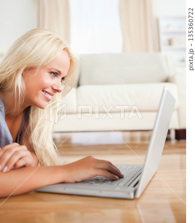 Blonde woman lying on floor typing on silver laptop beside light-colored sofa in living room Blonde woman lying on floor typing on silver laptop beside light-colored sofa in living room 135360722