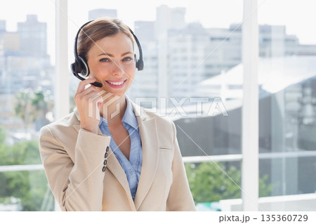 Mid adult woman standing in office wearing headset and smiling overlooking urban skyline via glass Mid adult woman standing in office wearing headset and smiling overlooking urban skyline via glass 135360729