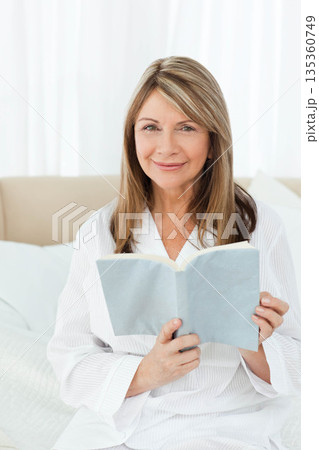 Senior woman reading light blue paperback book sitting on white bedding in bedroom with headboard 135360749