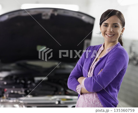 Woman standing with arms crossed in auto workshop beside open car hood and engine, copy space Woman standing with arms crossed in auto workshop beside open car hood and engine, copy space 135360759