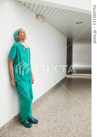 Woman leaning on hospital corridor wall wearing green scrubs scrub cap clogs mask, copy space Woman leaning on hospital corridor wall wearing green scrubs scrub cap clogs mask, copy space 135360762
