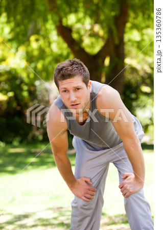 Man in grey workout clothes leaning with hands on knees catching breath in sunlit grassy park Man in grey workout clothes leaning with hands on knees catching breath in sunlit grassy park 135360786