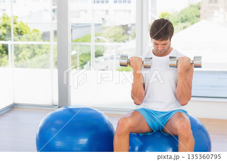Man in sportswear sitting on stability ball holding dumbbells at shoulder level at gym, copy space 135360795