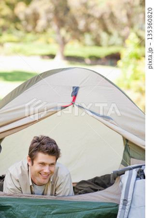 Mid adult man leaning from dome tent opening on sunny park campsite resting elbows beside backpack Mid adult man leaning from dome tent opening on sunny park campsite resting elbows beside backpack 135360820