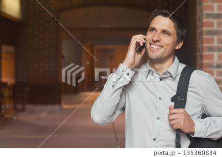 Mid adult man in grey shirt standing in brick corridor holding smartphone and backpack, copy space 135360834