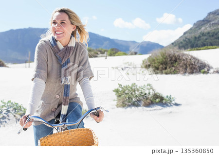 Mature woman riding bicycle across coastal sand dunes with wicker basket, copy space Mature woman riding bicycle across coastal sand dunes with wicker basket, copy space 135360850