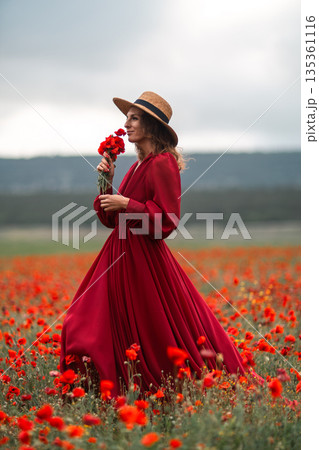 Woman poppy field. Elegant lady in a flowing red dress smelling flowers in a vast blooming meadow with copy space. Woman poppy field. Elegant lady in a flowing red dress smelling flowers in a vast blooming meadow with copy space. 135361116