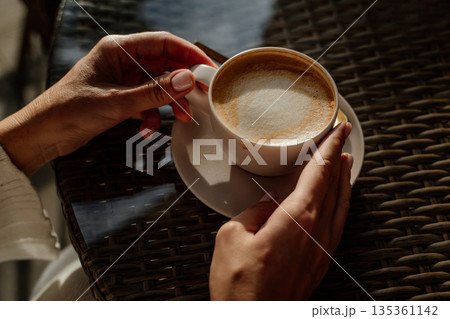 Coffee Cup Hands Table - Woman holds a cup of coffee with her hands on a woven table. 135361142