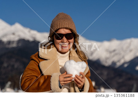 Snow Heart Mountains Winter: Woman holding snow heart, smiling, sunny day, snowy mountain backdrop. 135361188
