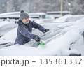 A man in a dark jacket and gray hat diligently removes a thick layer of fresh snow from the windshield of his car with a scraper and a broom. The entire scene takes place during a heavy snowfall in a 135361337