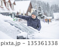 A man in a dark jacket and gray hat diligently removes a thick layer of fresh snow from the windshield of his car with a scraper and a broom. The entire scene takes place during a heavy snowfall in a 135361338