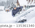 A man in a dark jacket and gray hat diligently removes a thick layer of fresh snow from the windshield of his car with a scraper and a broom. The entire scene takes place during a heavy snowfall in a 135361340
