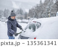 A man in a dark jacket and gray hat diligently removes a thick layer of fresh snow from the windshield of his car with a scraper and a broom. The entire scene takes place during a heavy snowfall in a 135361341