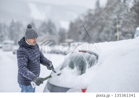 A man in a dark jacket and gray hat diligently removes a thick layer of fresh snow from the windshield of his car with a scraper and a broom. The entire scene takes place during a heavy snowfall in a 135361341