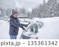 A man in a dark jacket and gray hat diligently removes a thick layer of fresh snow from the windshield of his car with a scraper and a broom. The entire scene takes place during a heavy snowfall in a 135361342