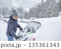 A man in a dark jacket and gray hat diligently removes a thick layer of fresh snow from the windshield of his car with a scraper and a broom. The entire scene takes place during a heavy snowfall in a 135361343